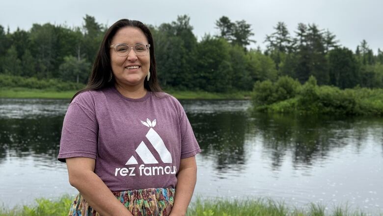 A woman with long brown hair wearing a purple shirt stands outside in front of a pond.