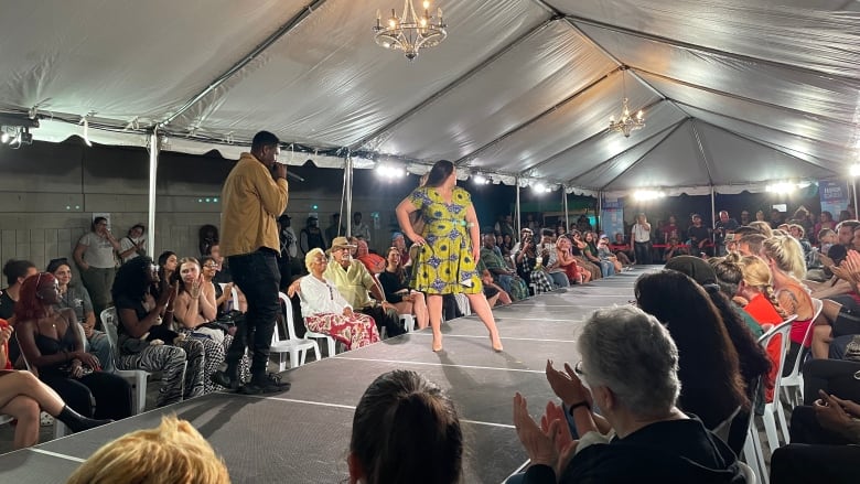 A woman in a dress walks a runway with people in chairs watching. 