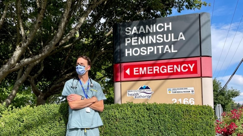 Doctor stands in blue scrubs with arms crossed wearing a stethoscope and mask in front of Saanich Peninsula Hospital.