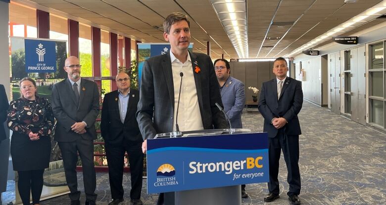 A man in a suit jacket and open-collared white shirt speaks at a podium bearing a sign that says StrongerBC.