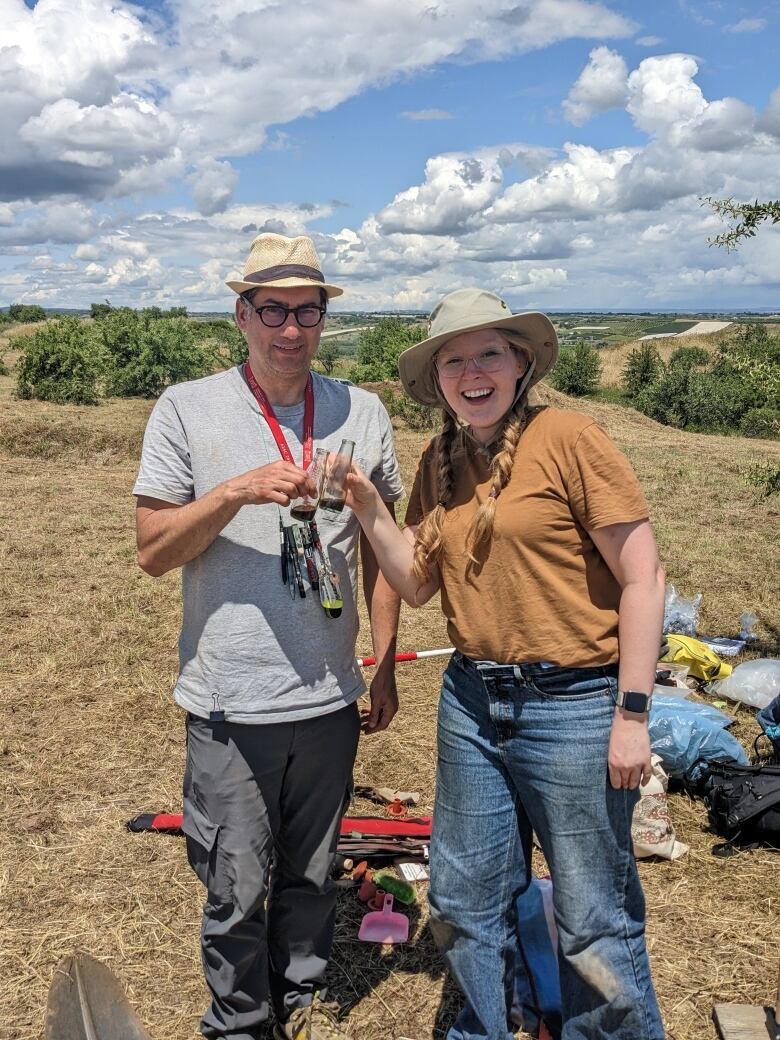 Male scientist and female colleague raise a glass of espresso with agricultural land in background