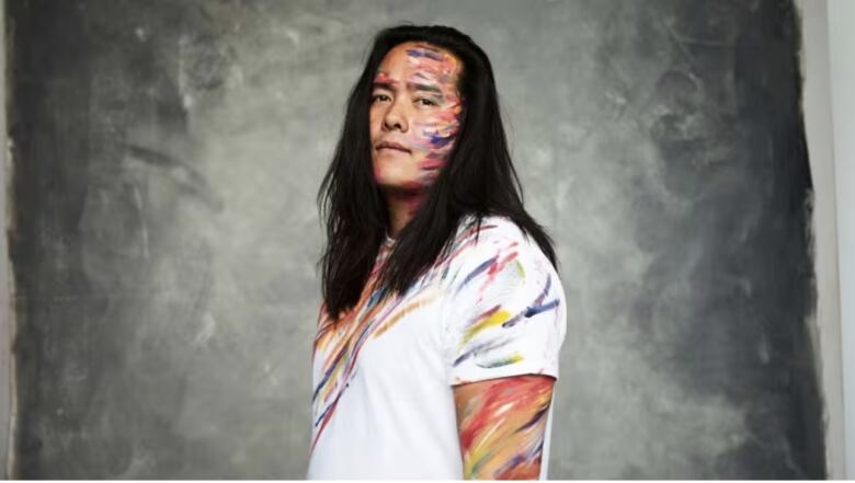 A headshot of Aysanabee - a man with long black hair wearing a white tshirt with colourful painstrokes on his shirt and face.