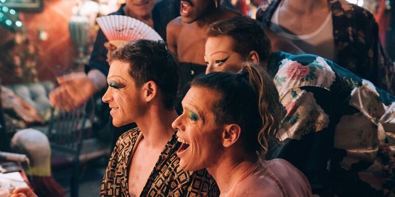 A group of smiling drag queens are shown looking into a mirror, as they prepare for a show. 