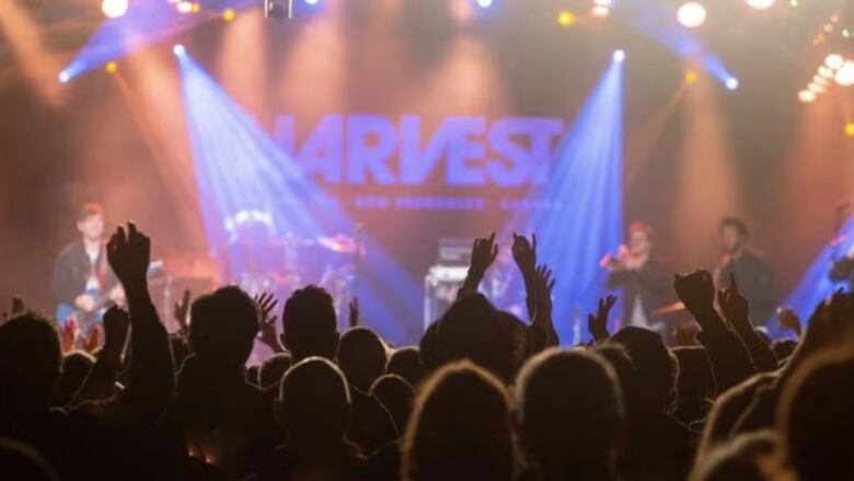 A stage lit with purple light, silhouettes of people with arms up in the foreground.