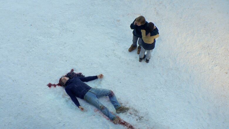 A body lies on its back in the snow, with blood pooling around the head. Nearby, a woman and young boy hold each other. 