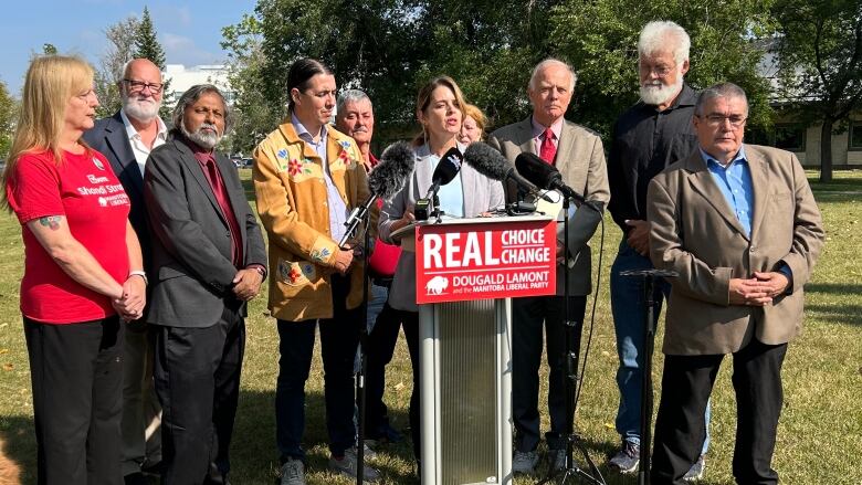 Woman stands behind podium speaking to media. A red Manitoba Liberal campaign sign hangs on the podium. The woman is surrounded by several other members of the party. 