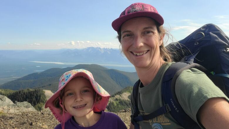 A woman with a backpack is seen smiling next to a girl wearing a pink hat and a purple T-shirt against the backdrop of the mountains.