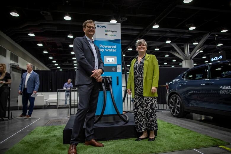 A man in a suit and a woman in a pantsuit and jacket stand on either side of a blue charging station that says, Powered by Water.