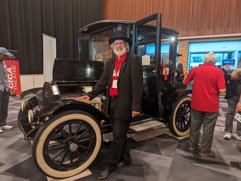 A man in a white beard and round-brimmed hat wearing a red sweater with a dark suit jacket over it stands next to an old vehicle with round spoked tires that looks a bit like a Model T but isn't.