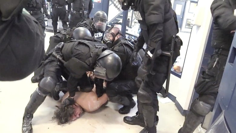 A shirtless man is held down on the floor by several people dressed in black riot gear.