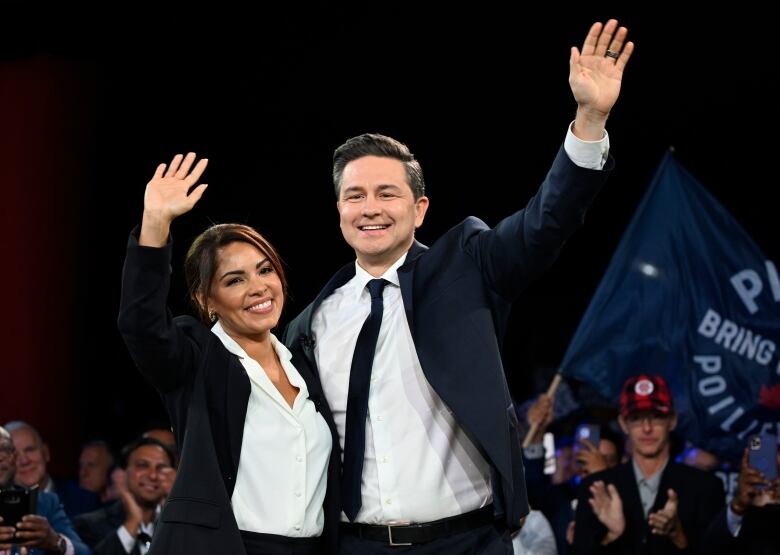 A man and a woman in business attire wave to a crowd on a convention hall stage.