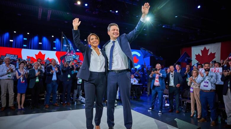 A man and a woman in business attire wave to a crowd on a convention hall stage.