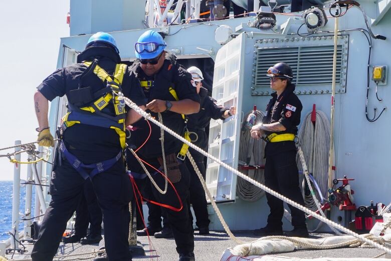 Two men on a ship deck, with one attaching safety lines to the other. In the background, another man is coming through a door while a fourth looks on. 