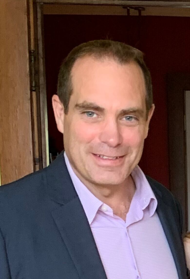 A headshot of a man with short brown hair wearing a dark suit jacket over a purple dress shirt.