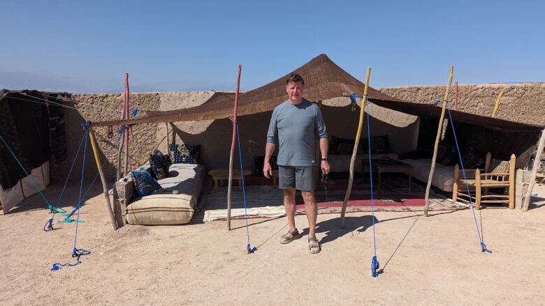 A man wearing a blue shirt stands in sand in front of a brown tent.