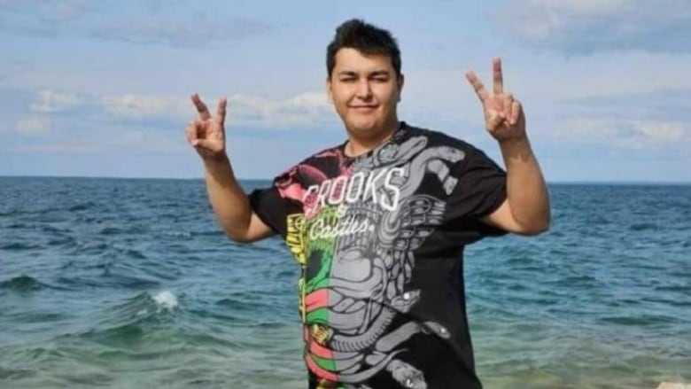 A young man stands next to a lake with two peace signs up in the air.