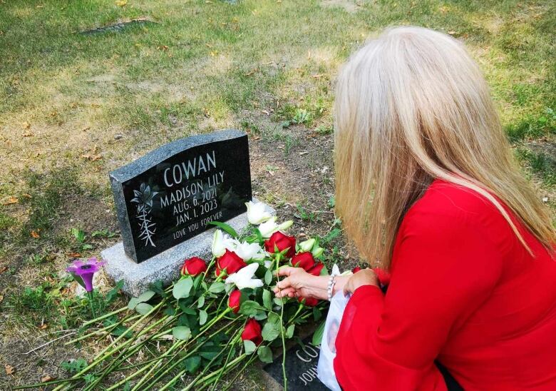 A blonde woman wearing a red jacket places roses at a tiny tombstone. The tombstone has Madison Cowan's name on it, and the dates she was born and died. 