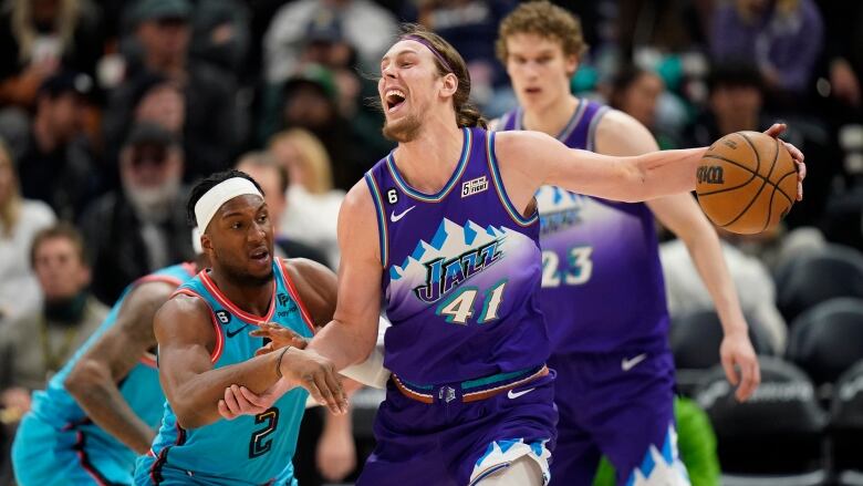 A man wearing a purple jersey holds a basketball away from a man wearing a blue jersey.