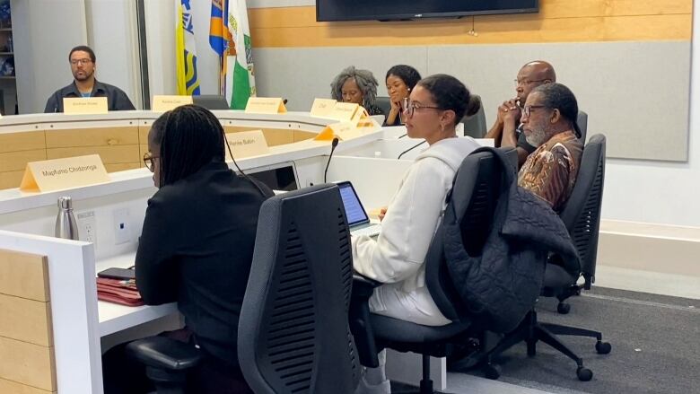 A group of seven Black people of various ages can be seen sitting around a horseshoe-shaped white table with microphones in front of them