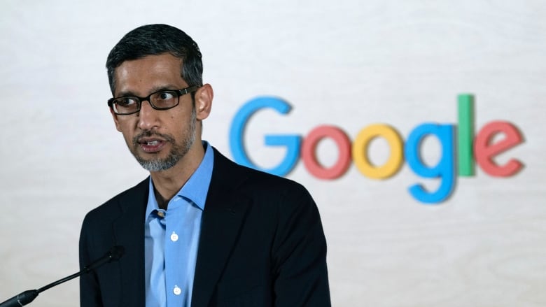 A bespectacled man in a suit jacket and collared shirt is shown speaking before a backdrop that says, 'Google.'