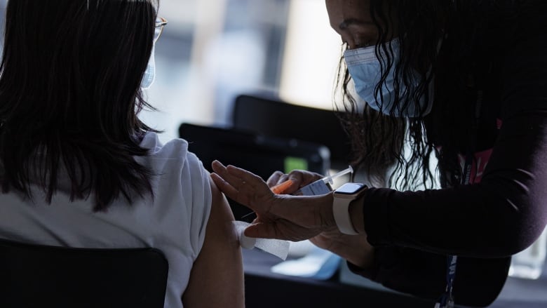 A nurse wearing a blue medical face mask pushes a needle into a young woman's upper arm.