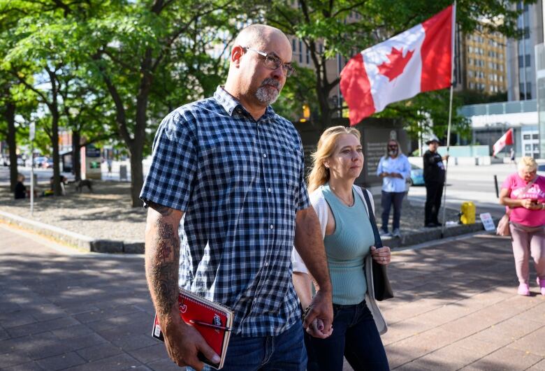 A man holds hands with a woman as they walk to a courthouse. Someone holds a Canada flag in the background.