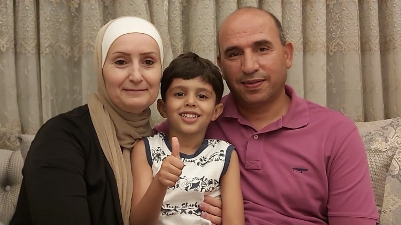 A family of three pose for a photo with a child between two parents. The child is giving a thumbs-up sign.