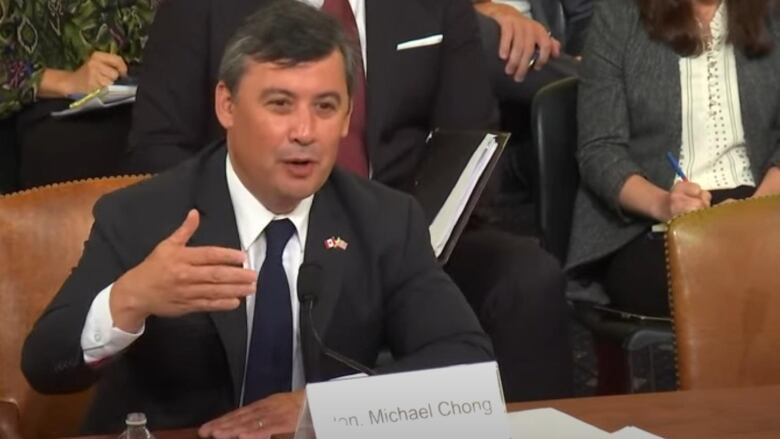 Chong sitting and gesturing while speaking, as people behind him listen or take notes.