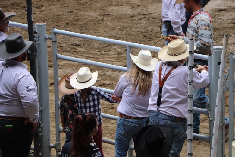 People wearing cowboy hats lean over a rail in a stadium looking at a rodeo.