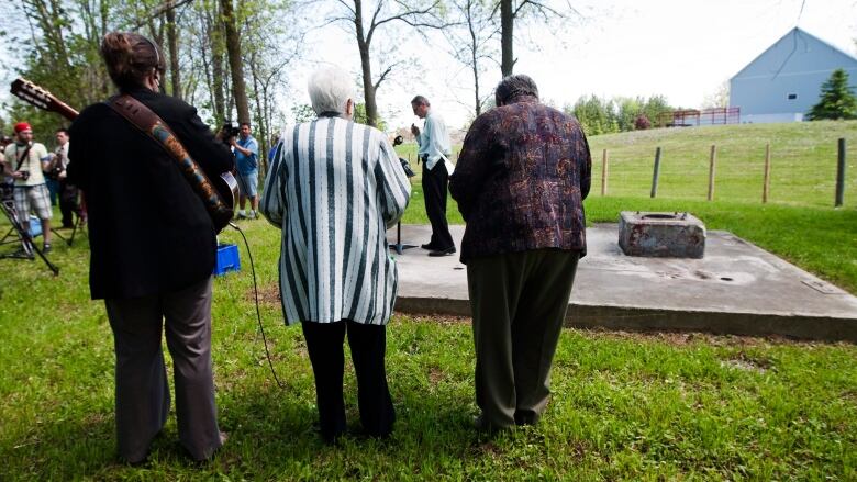 Three people stand with their backs to the camera watching a speaker who is standing on a closed well in Walkerton, Ont.