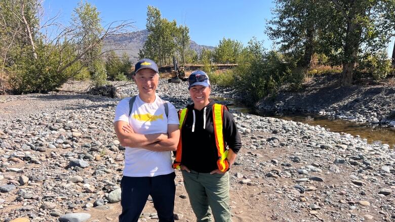 A middle-aged Asian man wears a white shirt and black jeans standing next to a woman wearing green cargo pants, a black hoodie and a safety vest. They are smiling on the banks of a creek in the sunshine.