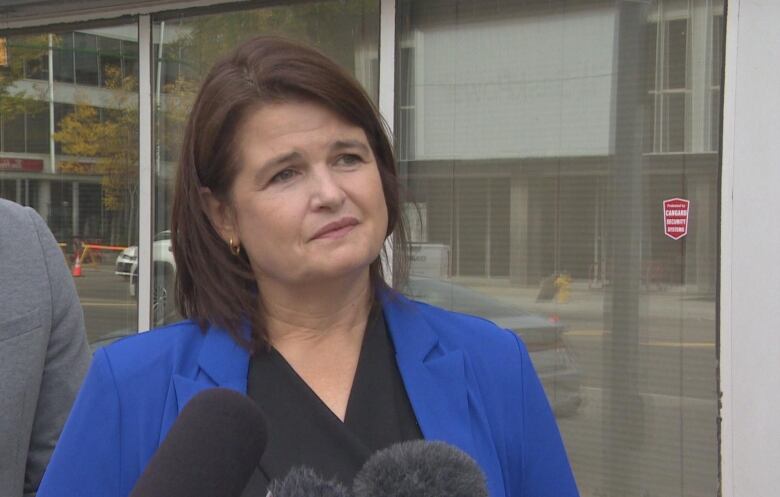 A woman with dark hair and a blue blazer stands in front of a building