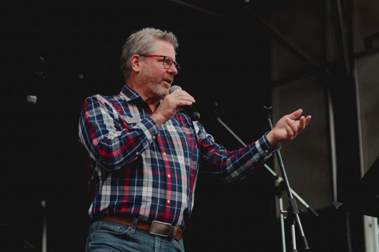 A white man with short grey hair, a beard and glasses stands on a stage in a red, white and blue plaid shirt and jeans, holding a microphone.