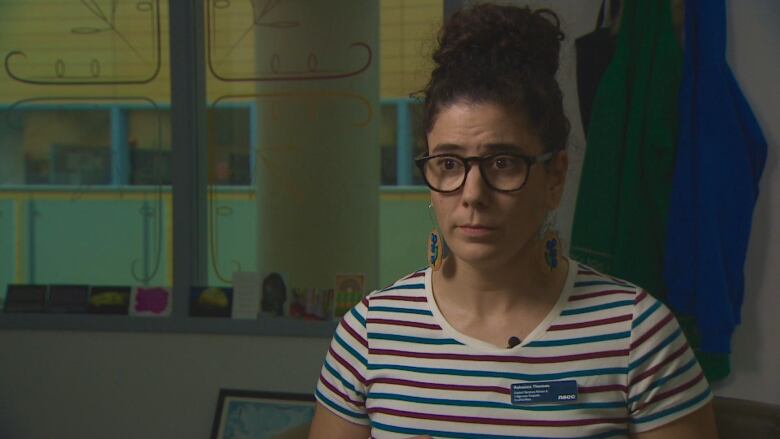 A woman has a concerned look on her face. She is wearing a white, red and blue T-shirt. She is also wearing black glasses, dangly earrings and her brown hair in a bun. 