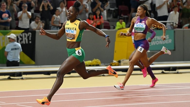 A female sprinter runs in the while two competitors are seen trailing behind her in the background during a race inside a track stadium filled with spectators.