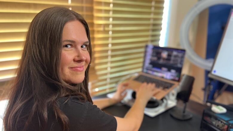 A woman with long brown hair sits in front of a laptop with her hands on the keyboard; she is looking at the camera and smiling.