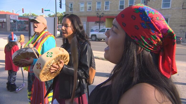 Three people sing and beat a traditional drum. A street lined with a building is behind them.