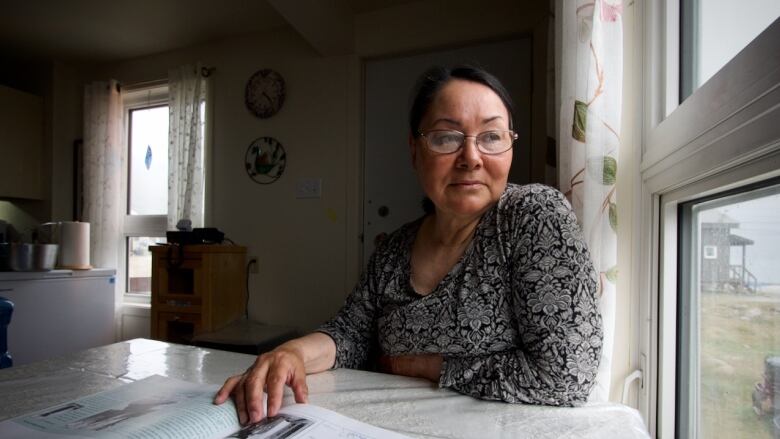 A woman sits at a table pointing to a photo in a book while looking wistfully out a window.