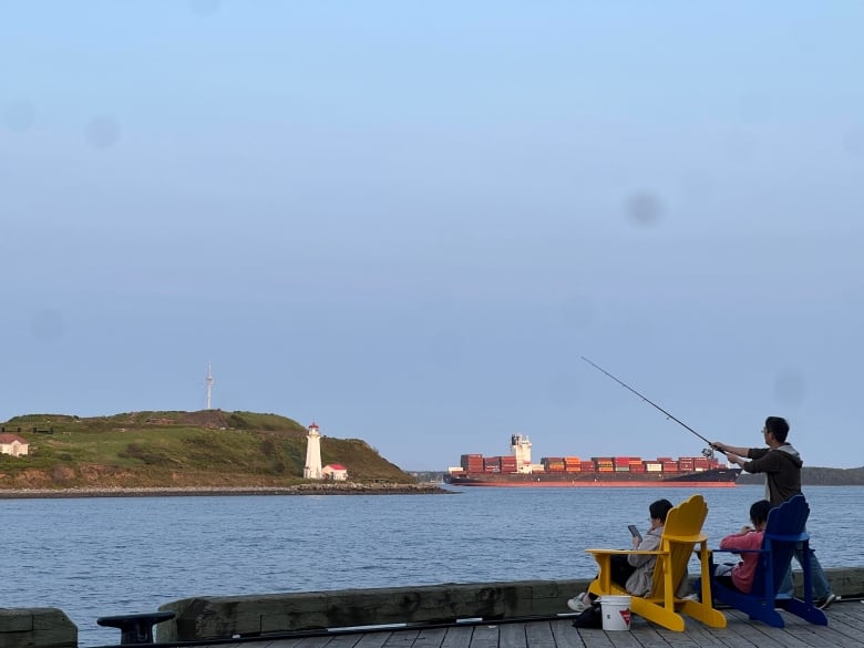 A man with a fishing rod casts his line beside a man sitting in a yellow adirondack chair. There is water, a lighthouse and container ship in the background.