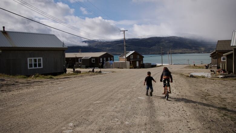 Two young children on a street photographed from behind with a view over the ocean. One is on foot. The other is on a bicycle. 