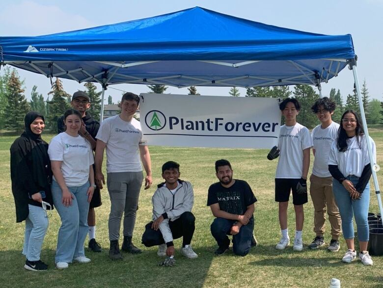 A group of young people stand in a park with a sign that reads Plant Forever.