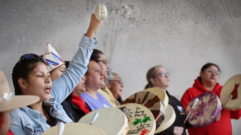 An Indigenous woman raises her mallet in the air while other women beat drums and sing.