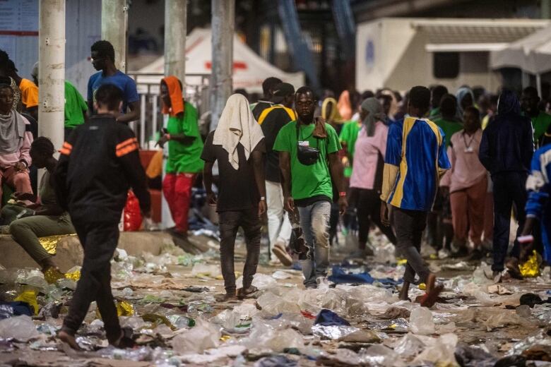 A crowd of people are shown at dusk, walking outside, with garbage and plastic littering the ground.