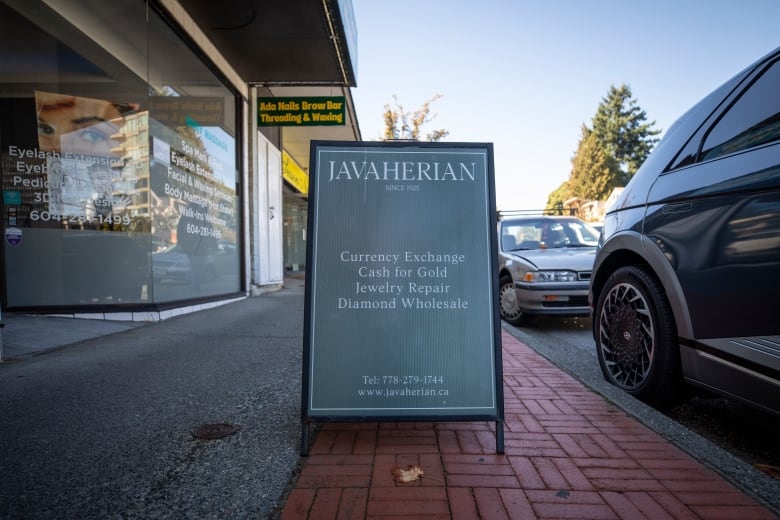 A forest green sandwich board sign advertising a jewelry consignment store is seen on a brick sidewalk on a sunny day.