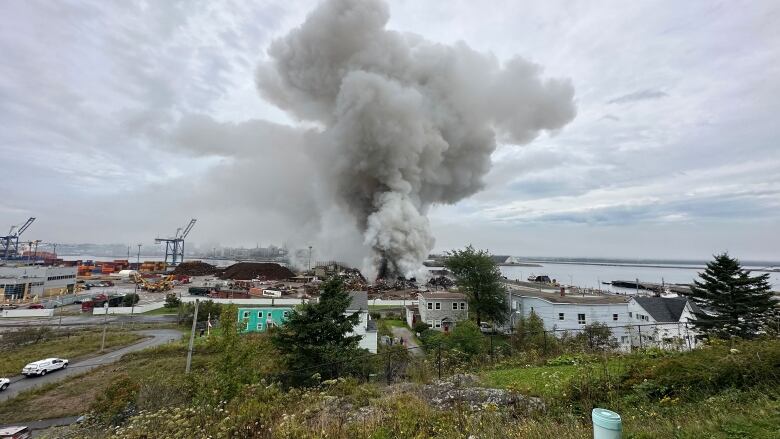 A large cloud of white and grey smoke hangs in the air above a fire at a scrap metal yard, surrounded by homes.