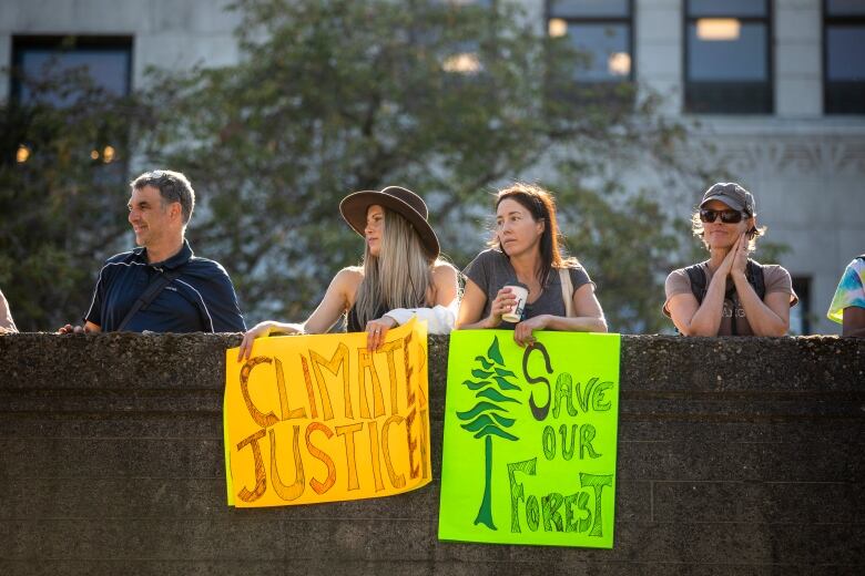 People are looking to the side at a concrete ledge. In the middle are two women holding an orange sign, reading 'Climate Justice,' and a green sign with a tree, reading 'Save Our Forest.'