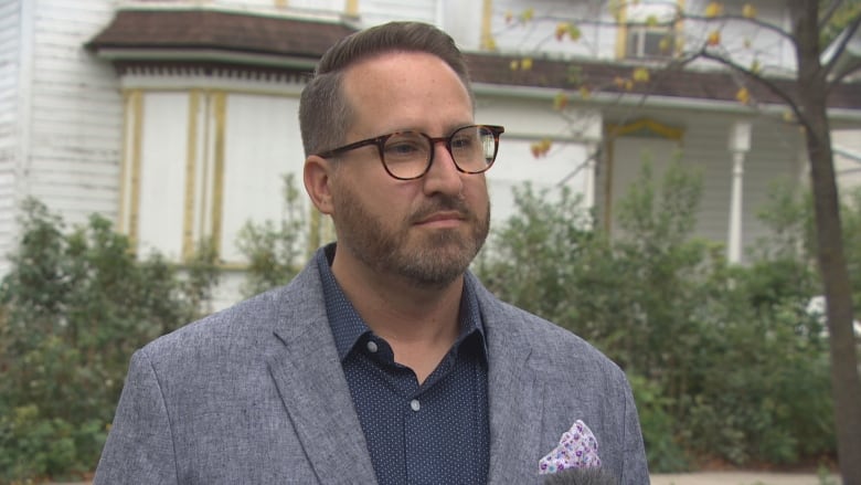 A man wearing a grey suit jacket stands in front of a white home.