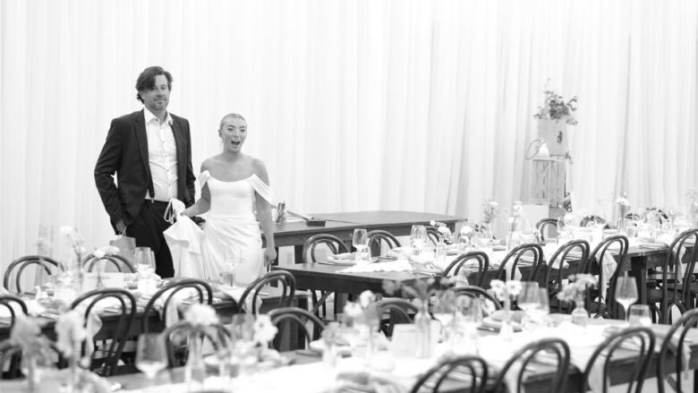 A bride and groom stand in a banquet room prepared for a wedding.