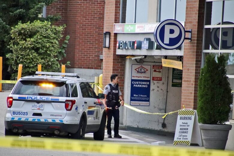 A parkade entrance is cordoned off by police tape and an officer stands outside it with an RCMP vehicle next to him.