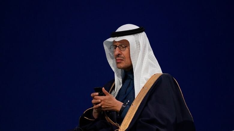 A man in an Arabian head dress holds his hands together and reads his phone as he waits his turn to speak